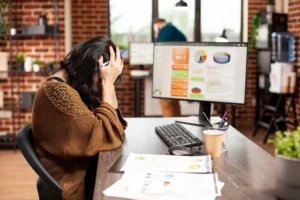 A woman sits at a desk with her head in her hands, appearing stressed. A computer displaying charts and papers is in front of her, in a modern office.